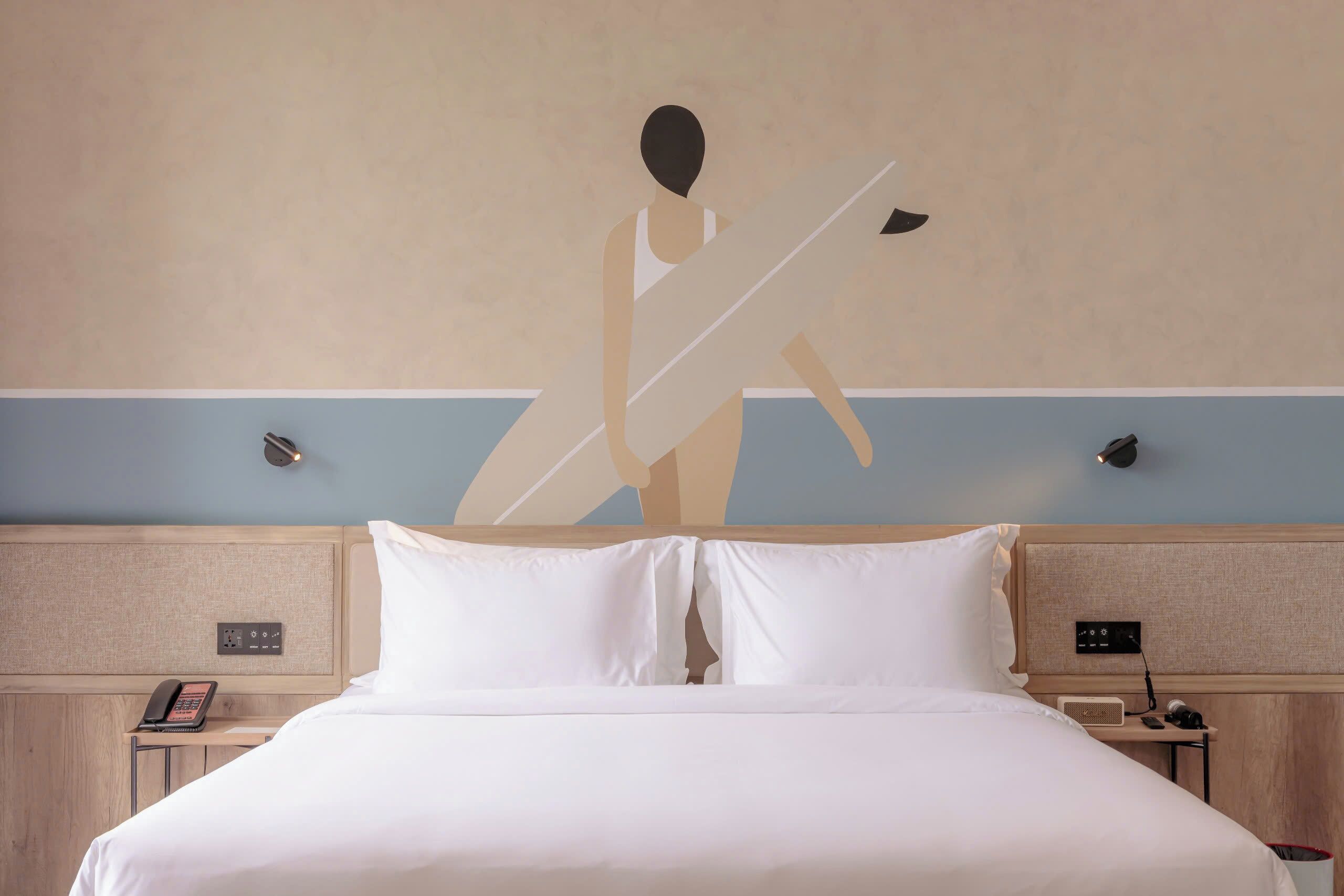 A hotel room with a bed, a nightstand with a phone and a radio, and a wall with a mural.