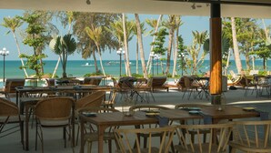 An outdoor dining area with tables and chairs, overlooking a beach with palm trees and the ocean.