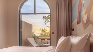 A hotel room with a large arched window, a bed with white linens, a small balcony with a table and chairs, and a painting on the wall.
