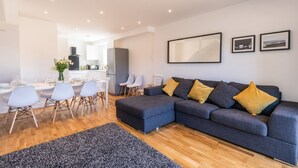 Un salon moderne avec un canapé d’angle gris, une table à manger avec des chaises, et une cuisine en arrière-plan.