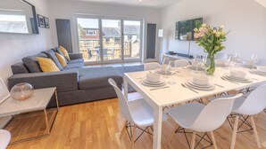 Un salon moderne avec un canapé gris, une table à manger blanche pour quatre personnes et une grande porte-fenêtre coulissante donnant sur un balcon.