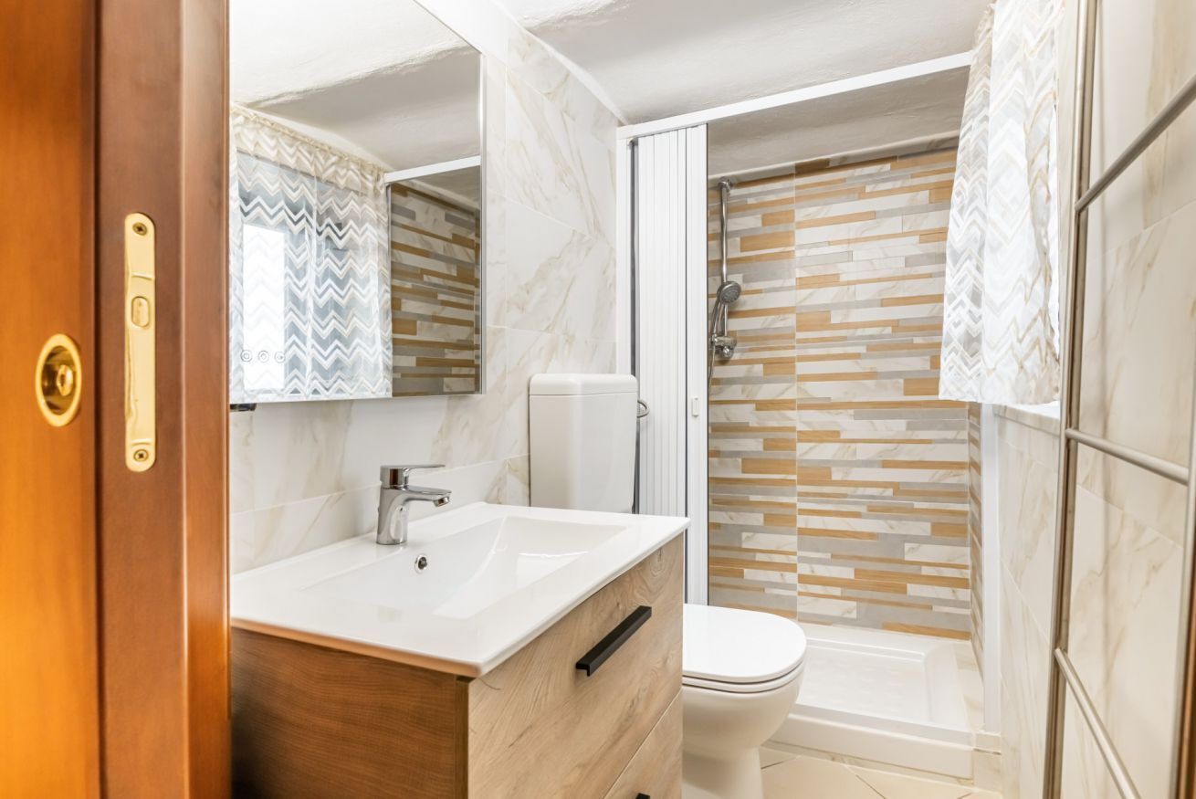 Casa de banho com toucador de madeira, lavatório branco, sanita e chuveiro com parede de azulejos estampados.