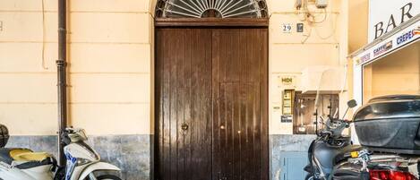 A closed dark wooden door with a decorative metal arch above it, a scooter parked to the left, and a motorcycle to the right.