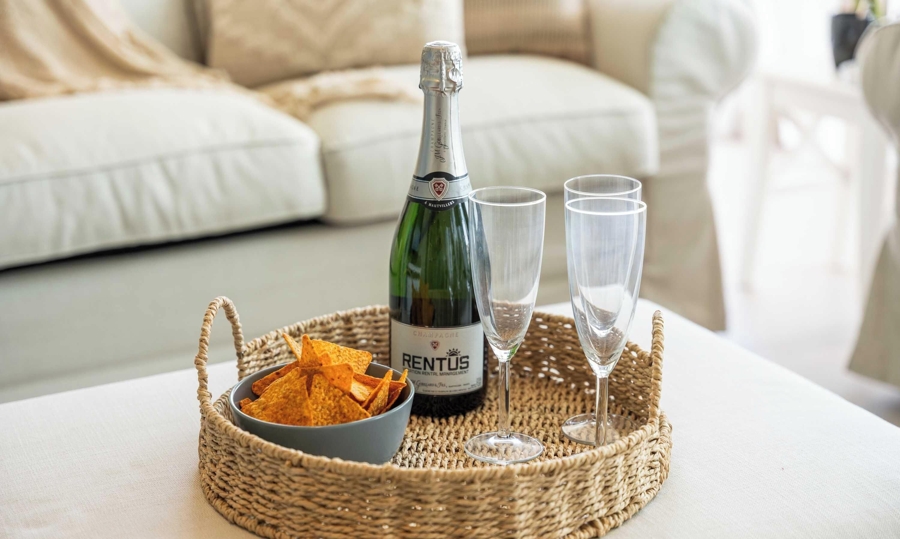 En flaska champagne, två glas och en skål med chips på ett flätat bricka.