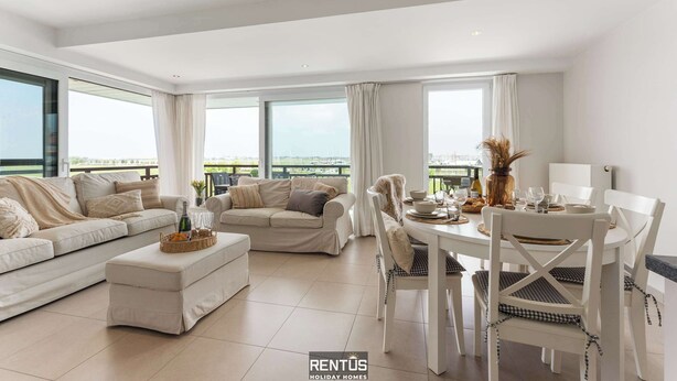 A spacious living room with a sofa set, a dining table with chairs, and large windows offering a view of the outdoors. - Seange - with beautiful view and many facilities (Nieuwpoort)
