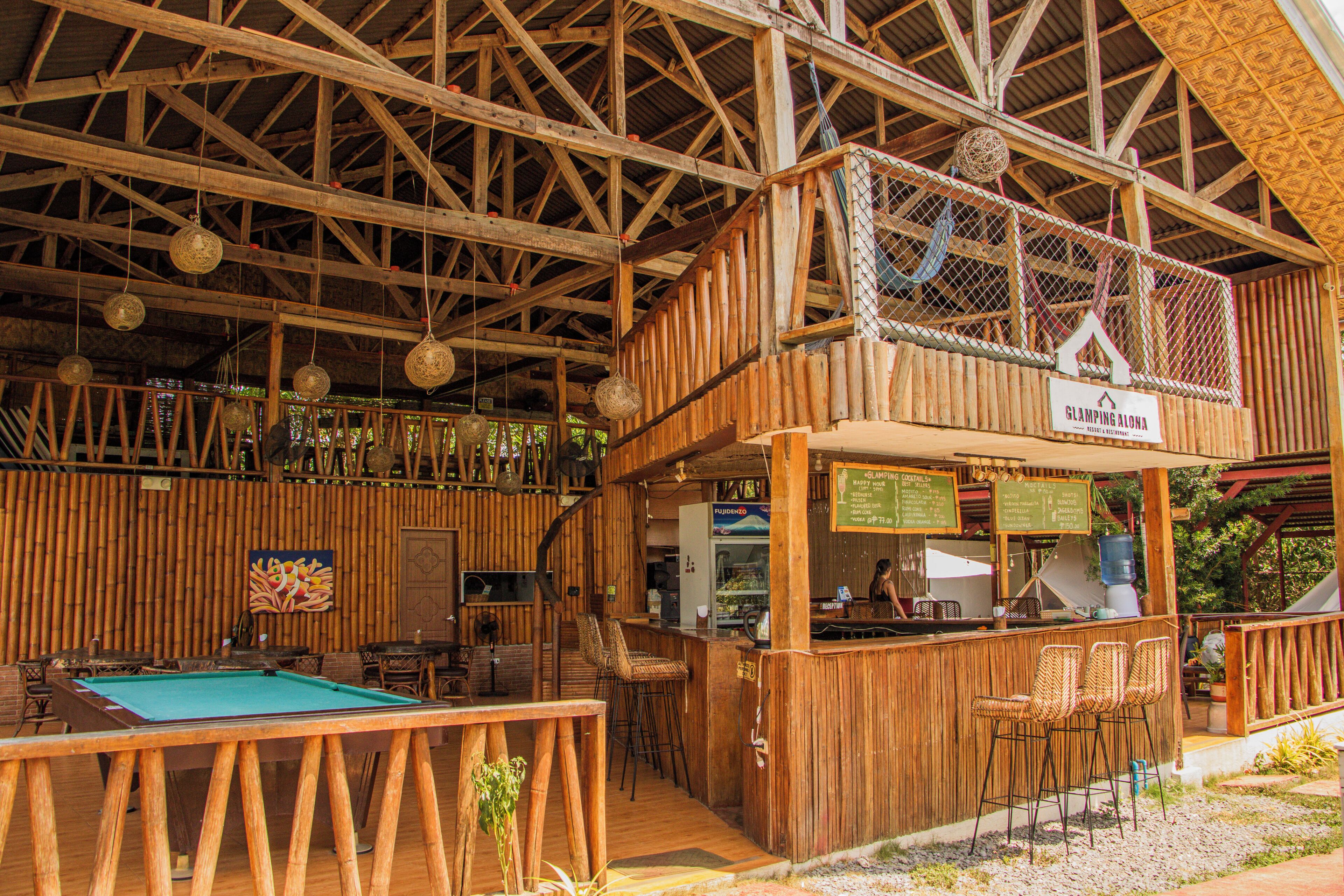 Zona de bar con barra de madera, mesa de billar, asientos y un letrero que dice Glamping Aloha.