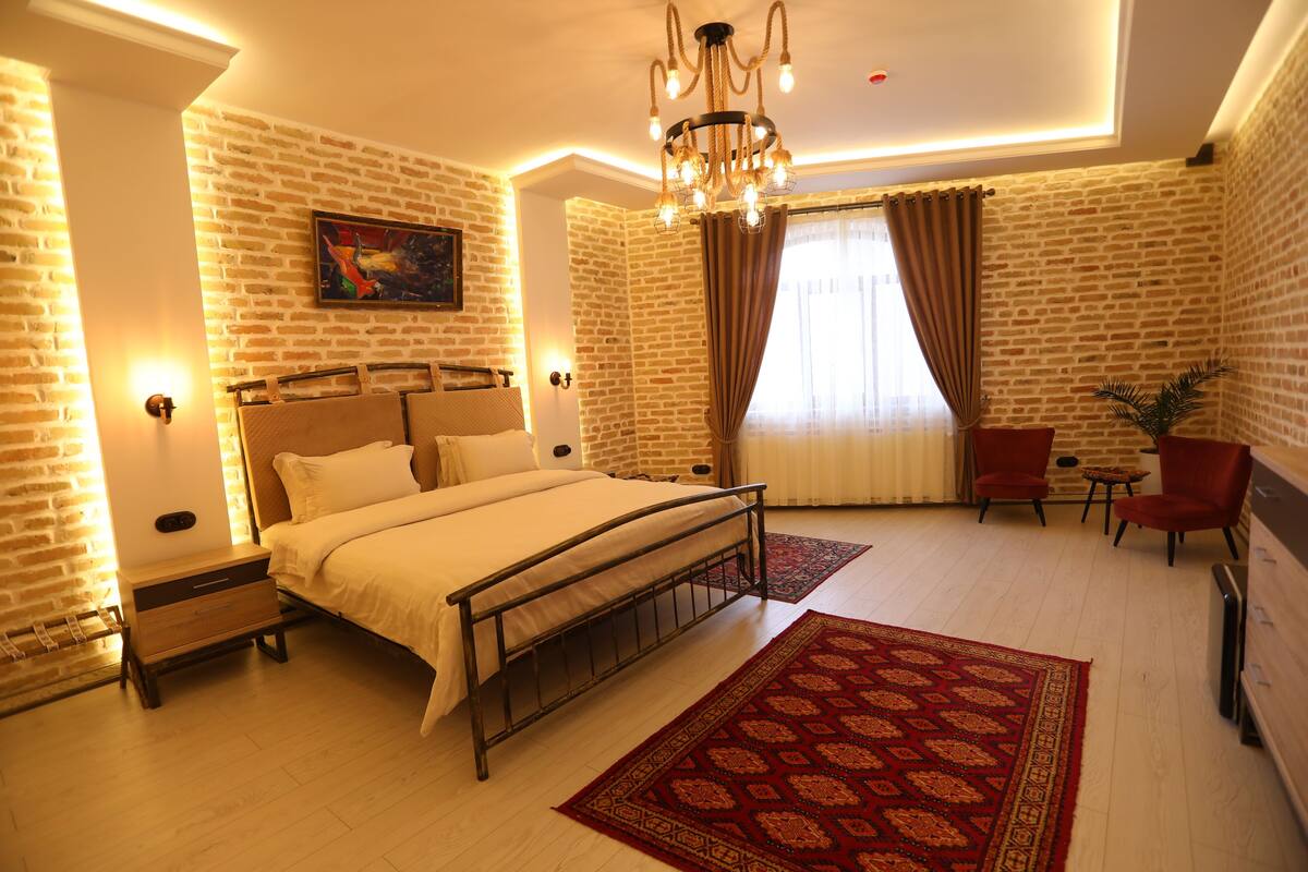 A bedroom with a brick wall, a bed with a metal frame, a chandelier, a red patterned rug, and a window with curtains.