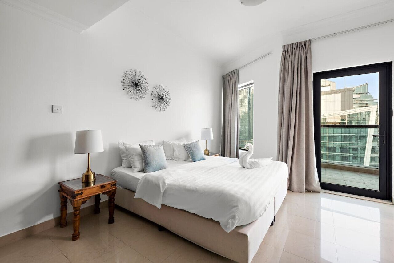 A modern bedroom with a large bed, two wall clocks, a bedside table with a lamp, and a view of the city through a large window.