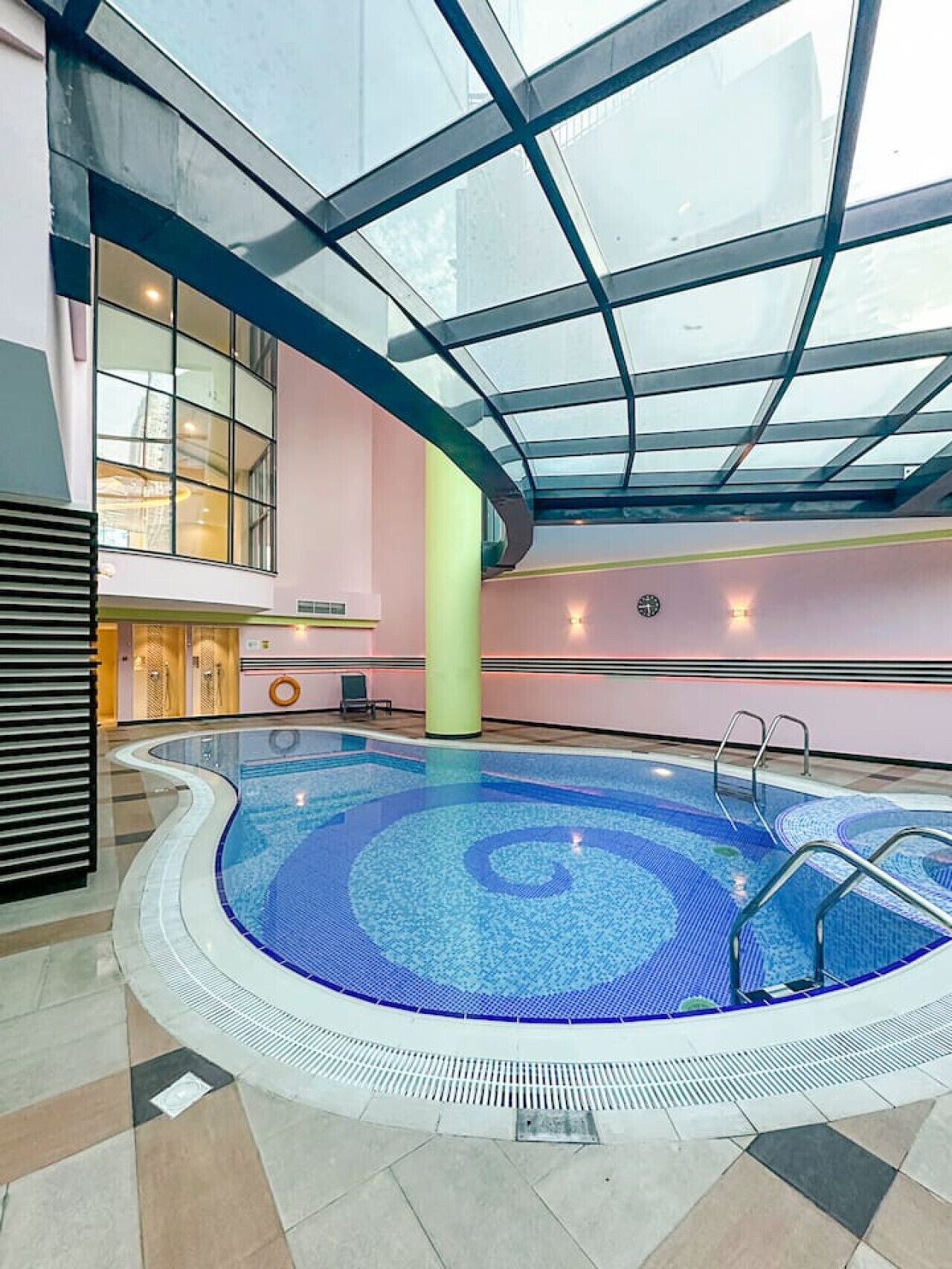 An indoor swimming pool with a glass ceiling, surrounded by a pink wall and a tiled floor.