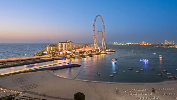 Eine Küstenstadt mit einem markanten Riesenrad, einem Hafen mit Booten und einer Strandpromenade mit Sonnenschirmen.