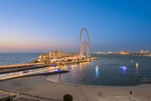 A coastal cityscape with a prominent Ferris wheel, a marina with boats, and a beachfront area with umbrellas. - World Class | 2 BR | Vibrant Waterfront District (JBR)