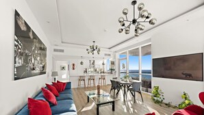 Ein modernes Wohnzimmer mit einer Couch, einem Couchtisch und einem Essbereich mit Stühlen. Ein großes Fenster bietet Blick auf das Meer.