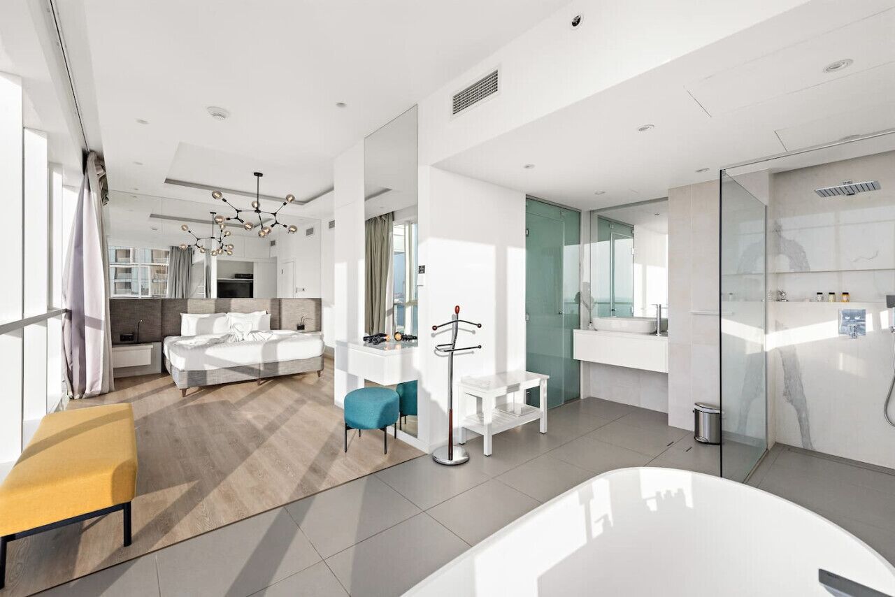 Una habitación de hotel moderna con una cama grande, una zona de estar, una mesita pequeña, un perchero y un baño con cabina de ducha de cristal.