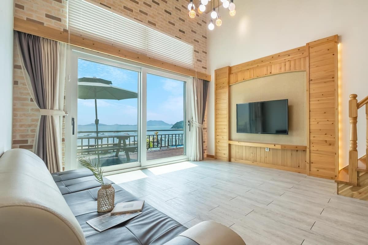 A spacious living room with a large sliding glass door leading to a balcony, a flat-screen TV mounted on the wall, and a wooden staircase.