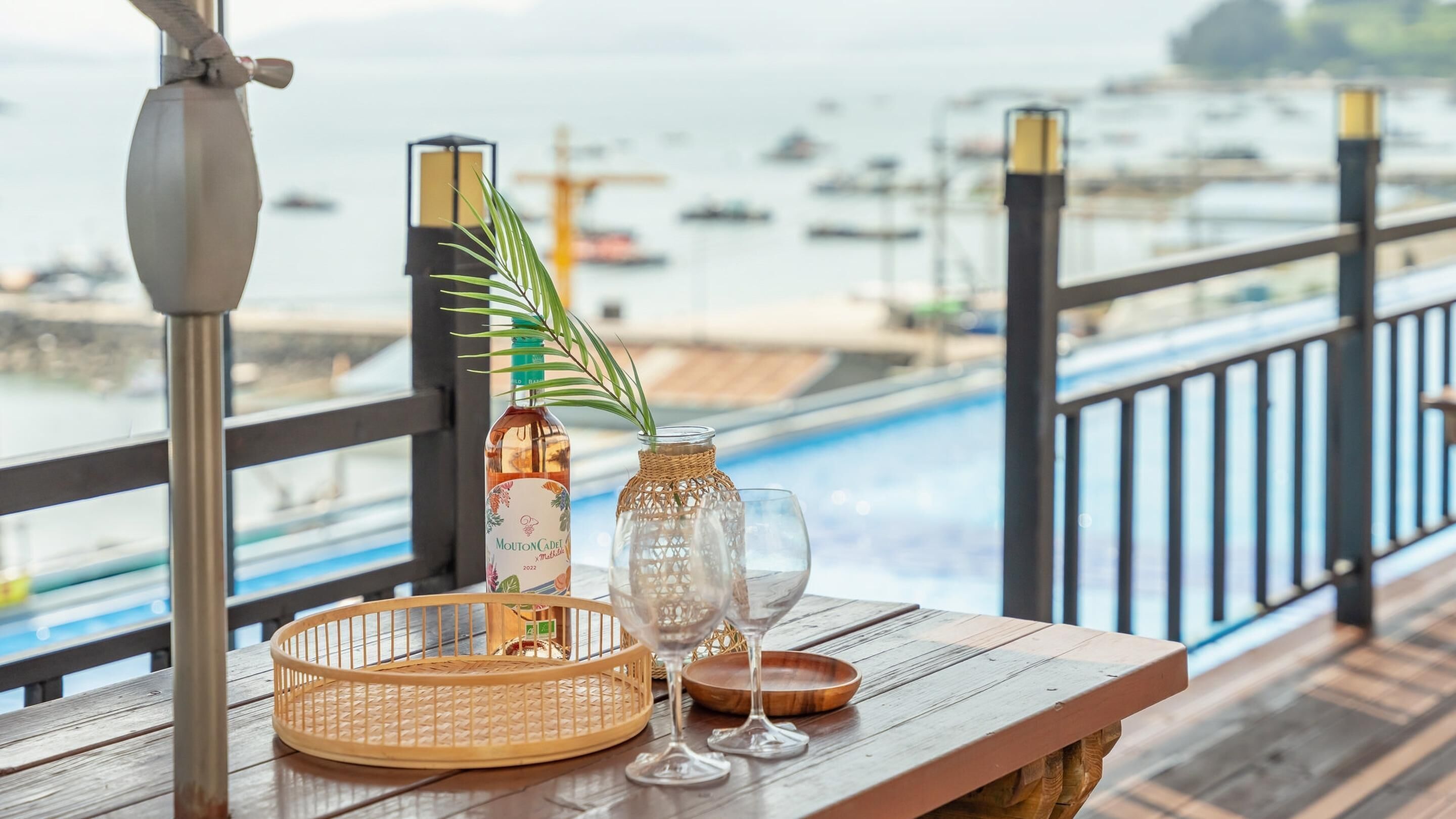 Una mesa de madera con una botella de Mouton Cadre, un florero con una planta y dos copas de vino en un balcón con vistas a un puerto deportivo lleno de barcos.