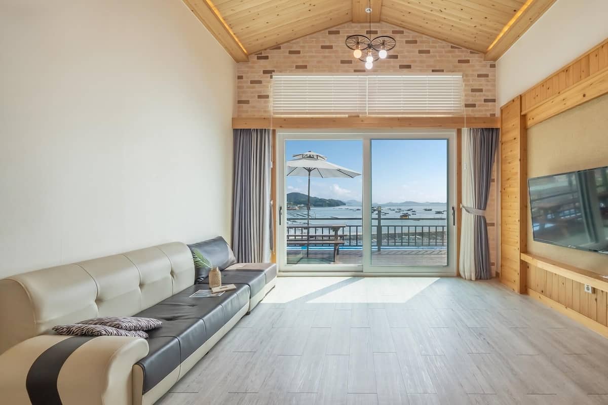 A spacious living room with a large TV, a sofa, and a view of the sea through a sliding glass door.