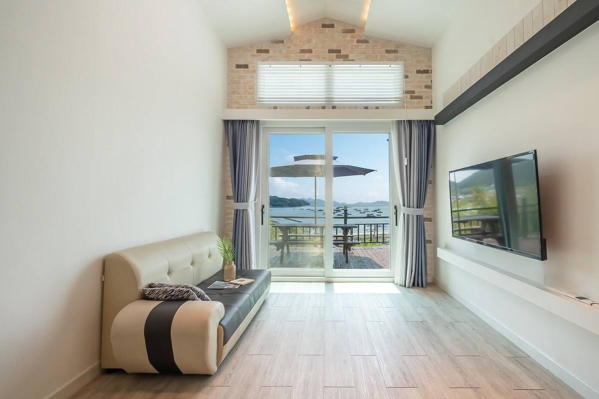 A modern living room with a sofa, a TV, and a view of the sea through a glass door.