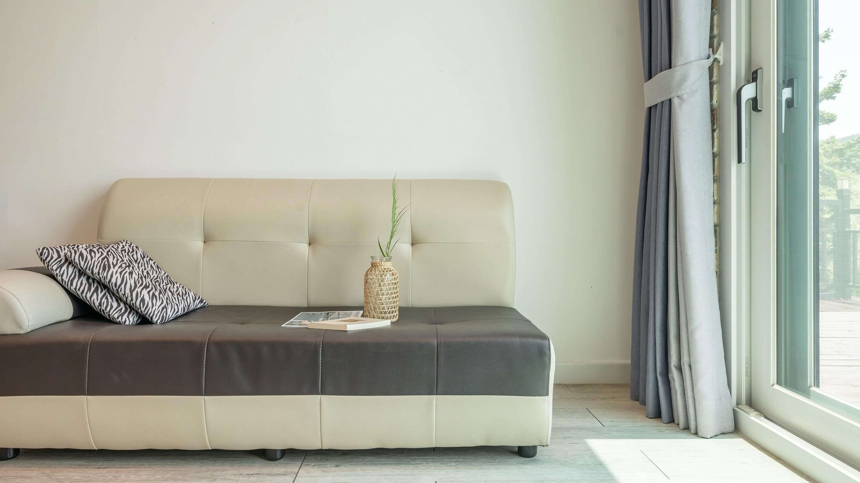 Una sala de estar moderna con un sofá beige, un cojín estampado, un florero de vidrio con una planta y una ventana con cortinas.