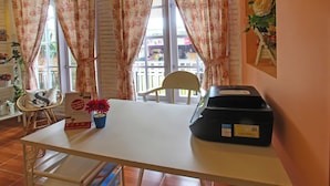 Une pièce avec une table blanche, une chaise, un bureau avec un ordinateur, une petite plante en pot et une fenêtre avec des rideaux.