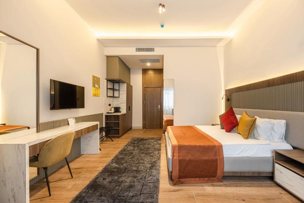 Una habitación de hotel moderna con cama, mesita de noche y escritorio con silla. Hay un televisor montado en la pared y una pequeña zona de kitchenette.