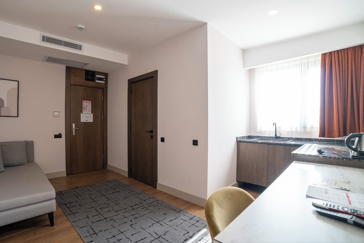 Una habitación de hotel moderna con sofá, un escritorio pequeño, una ventana con cortinas y una zona de cocina.