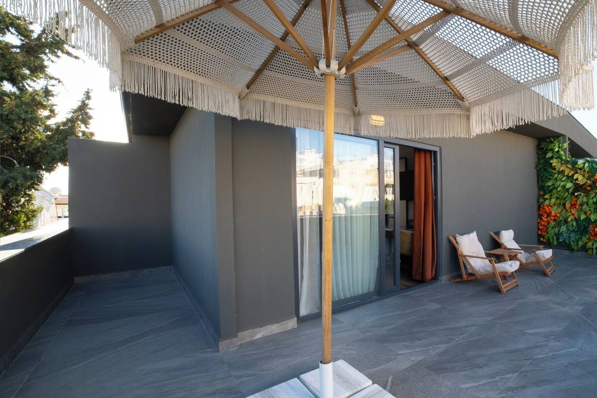 Un patio exterior moderno con un amplio parasol tejido, dos sillones de madera y una pared decorativa con follaje.
