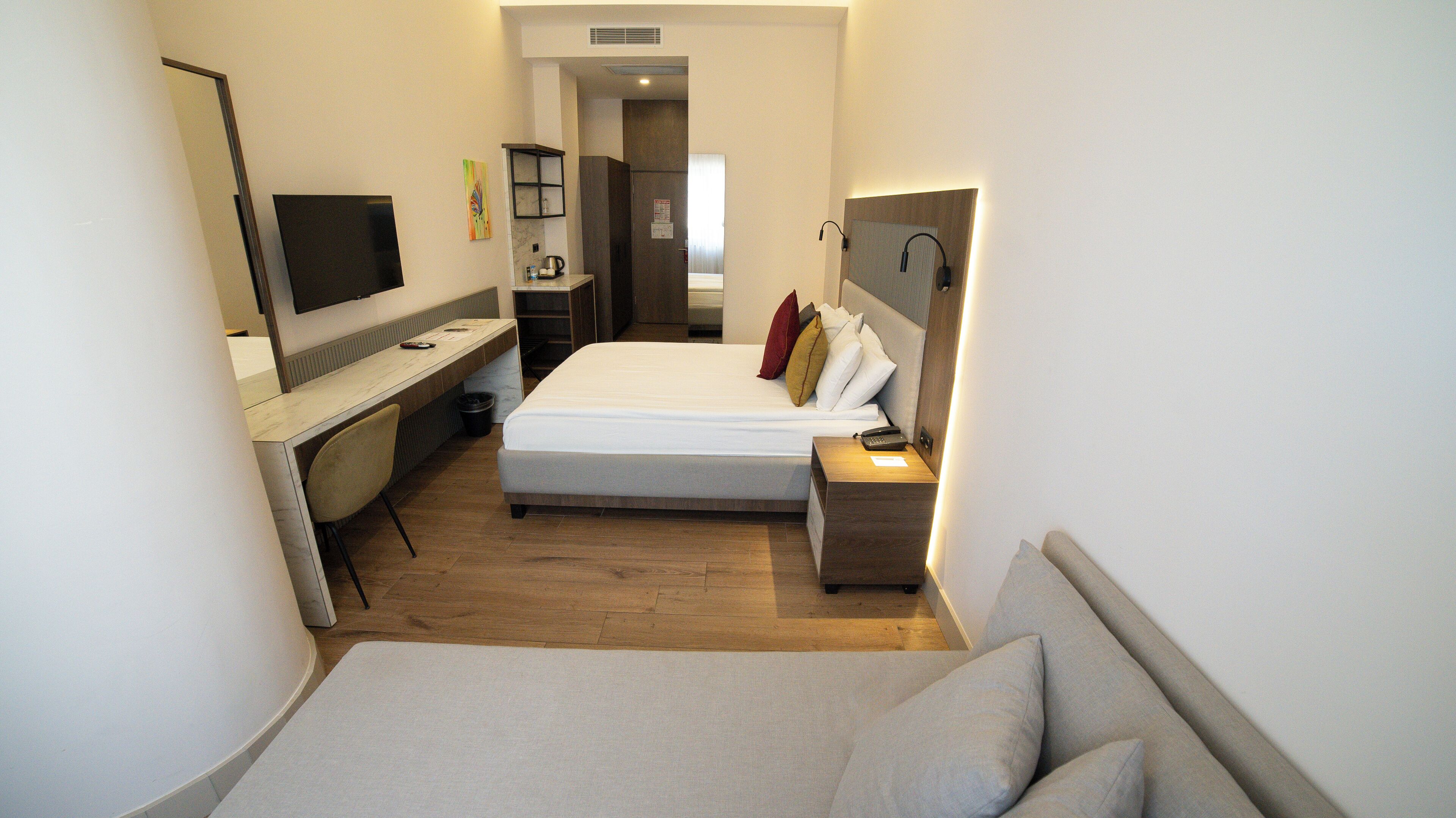 A compact hotel room with a bed, a desk, a chair, a TV, and a bedside table with a lamp.
