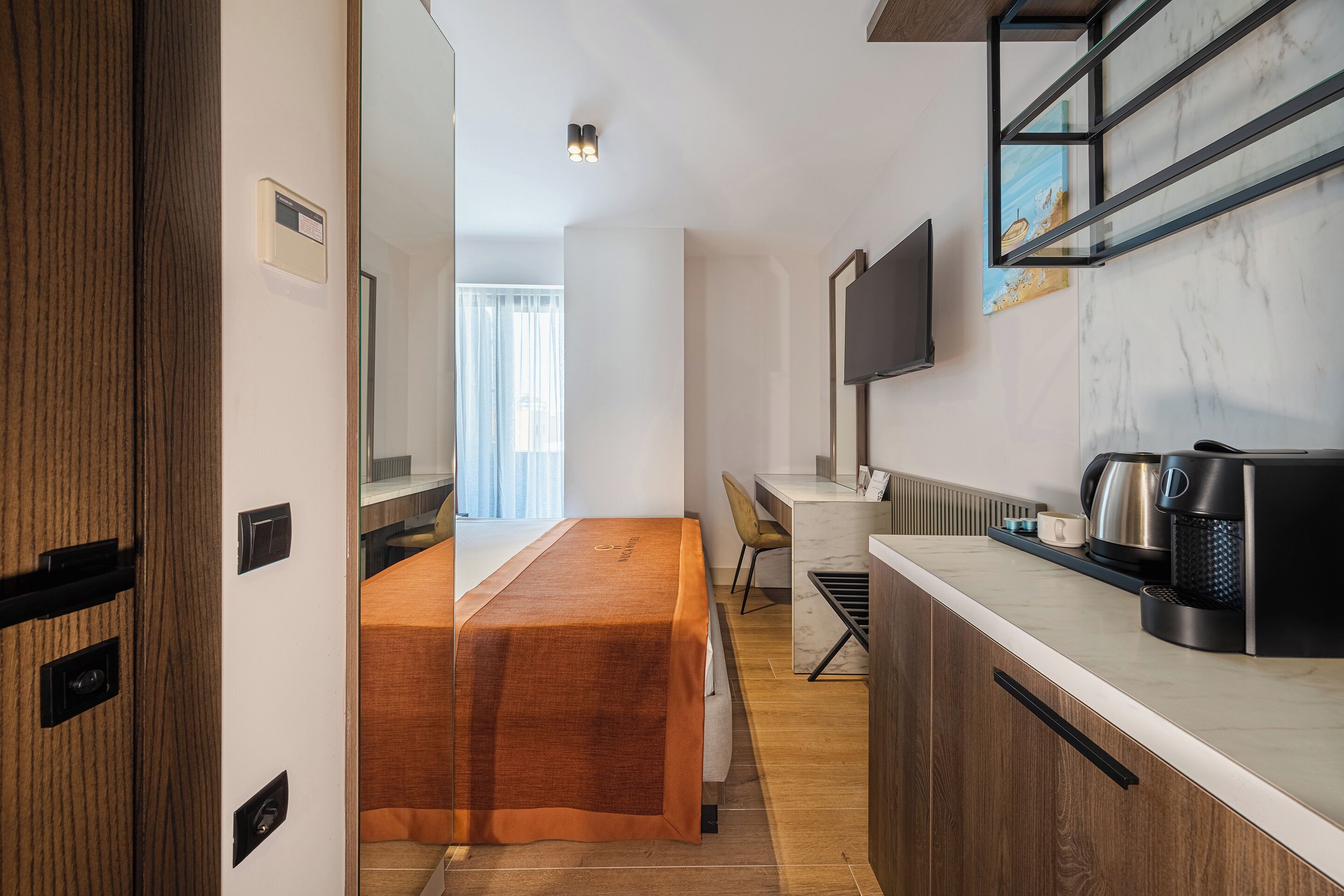 Ein kleines Hotelzimmer mit Bett, Schreibtisch, Kaffeemaschine und einem an der Wand montierten Fernseher.