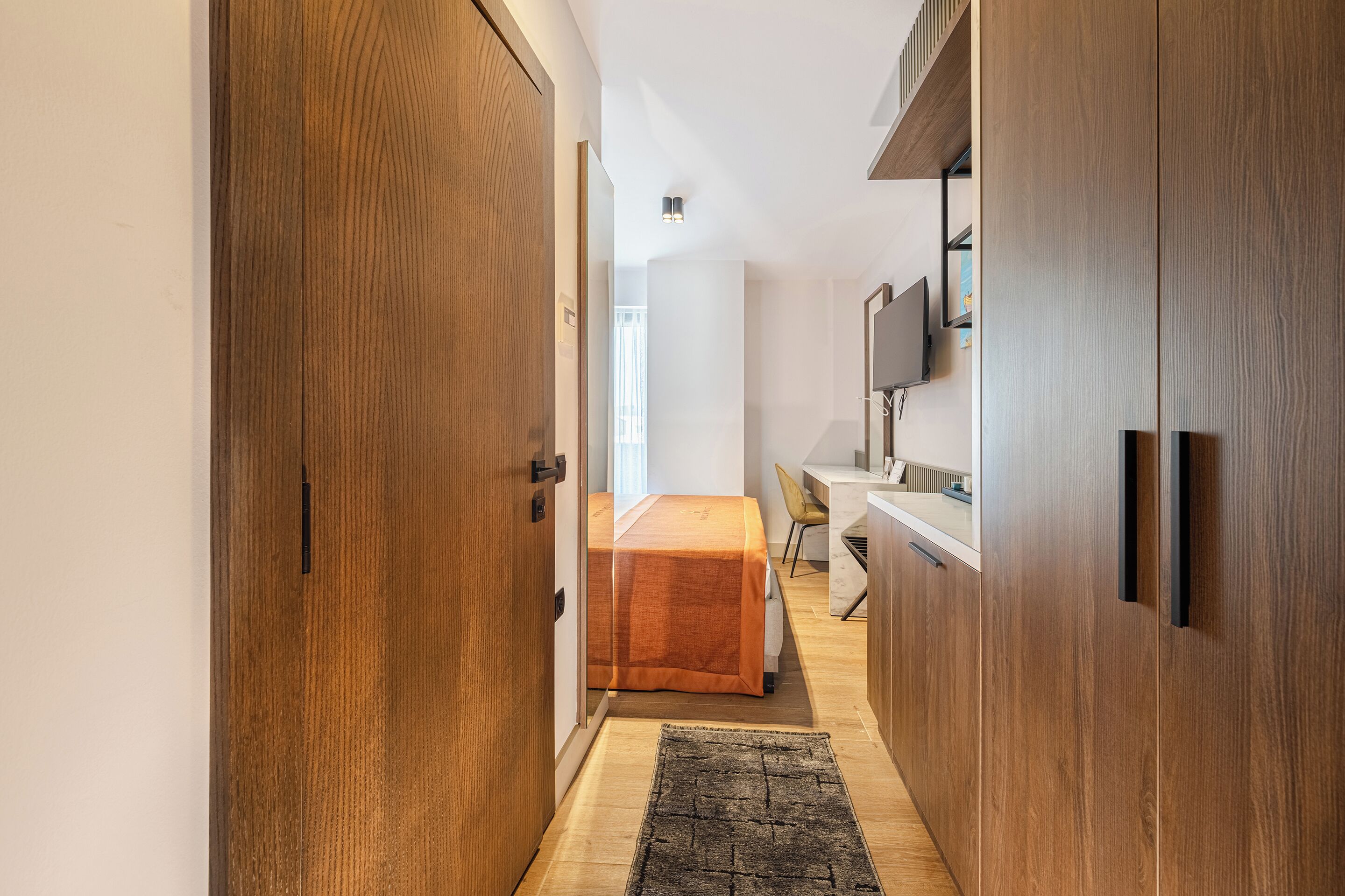 A narrow hallway with wooden doors, a bed with an orange bedspread, a desk with a chair, and a mounted TV on the wall.