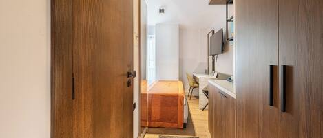 A narrow hallway with wooden doors, a bed with an orange bedspread, a desk with a chair, and a mounted TV on the wall.