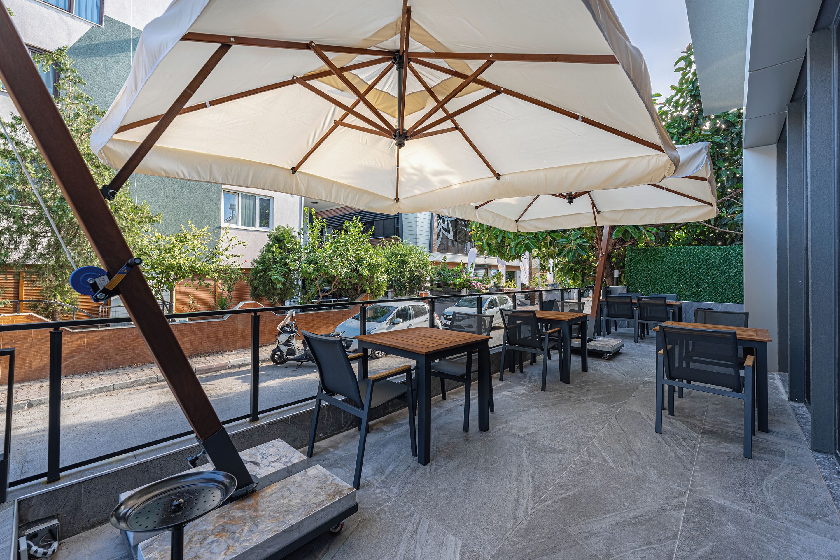 An outdoor seating area with tables and chairs under large umbrellas, a metal railing, and a view of the street and buildings in the background.