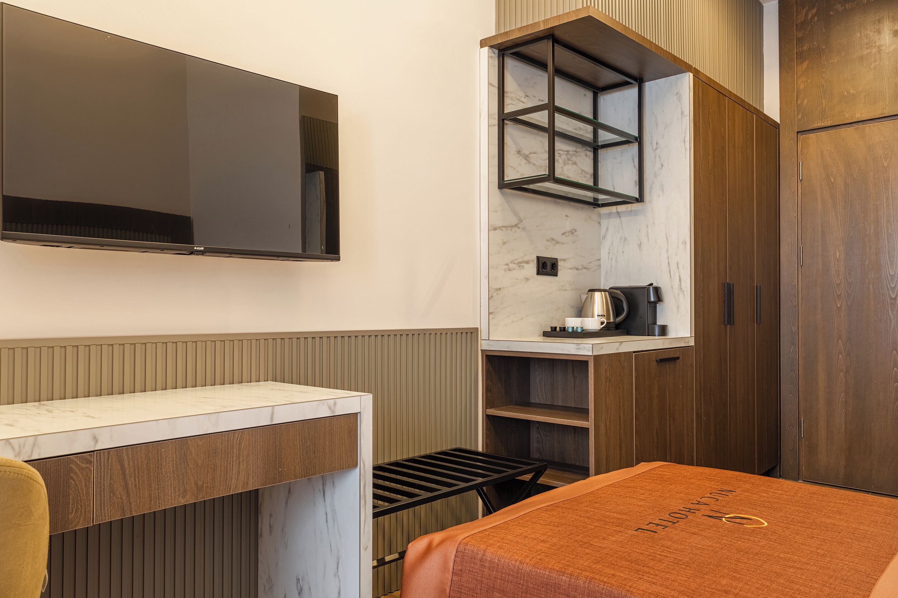A compact hotel room with a bed, a TV mounted on the wall, a small kitchen area, and a wardrobe.