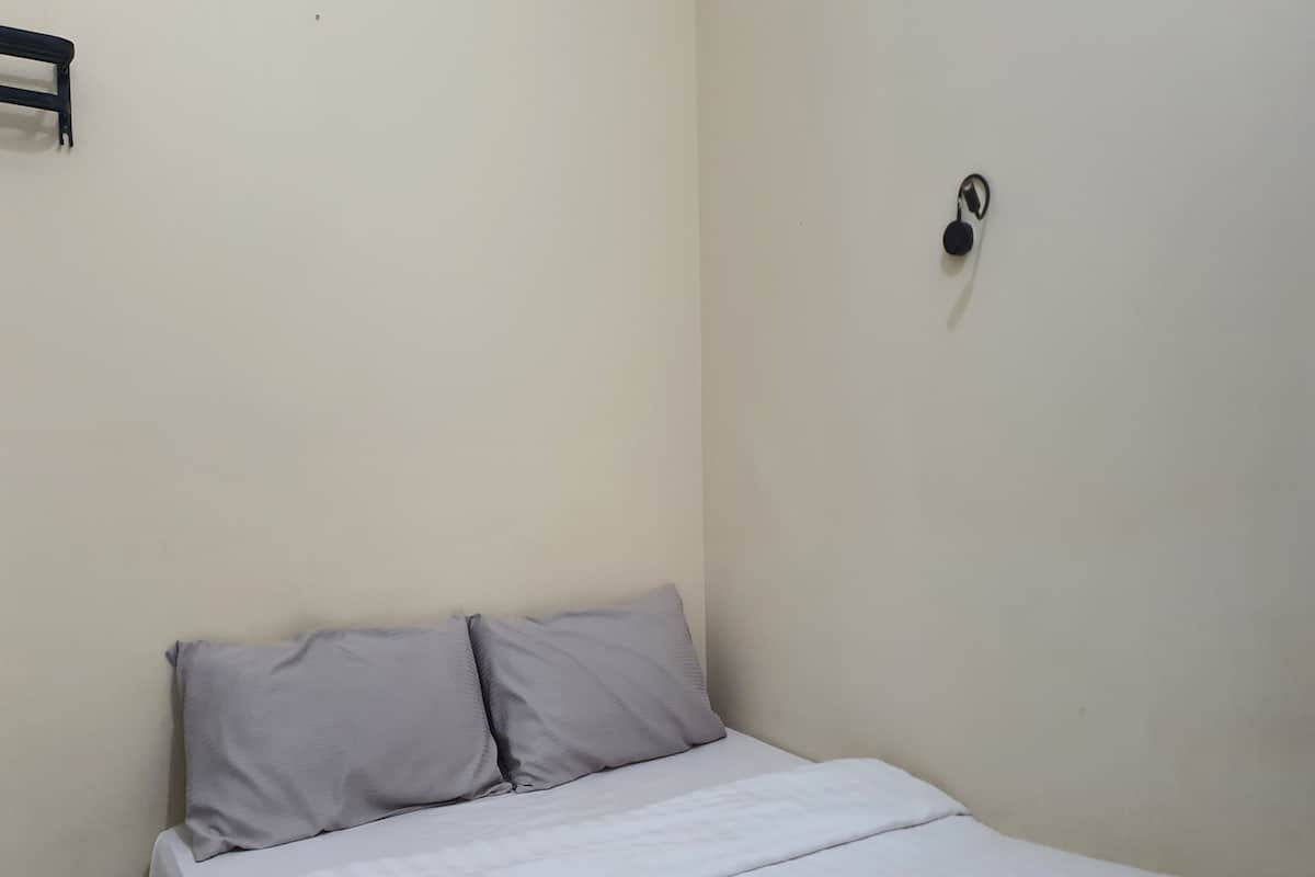 A small, single-bed room with a plain white bedspread and two pillows. There is a small black object on the wall to the right of the bed.