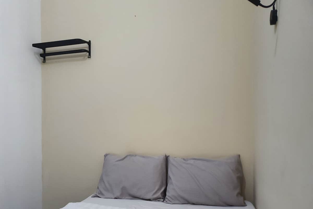A small, narrow room with a single bed, two pillows, and a wall-mounted shelf.