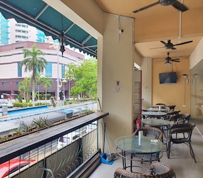 A balcony with a view of a building, a pool, and a street. There are wicker chairs and a table, and a ceiling fan. - SPOT ON 90984 Gn Homestay (Miri)