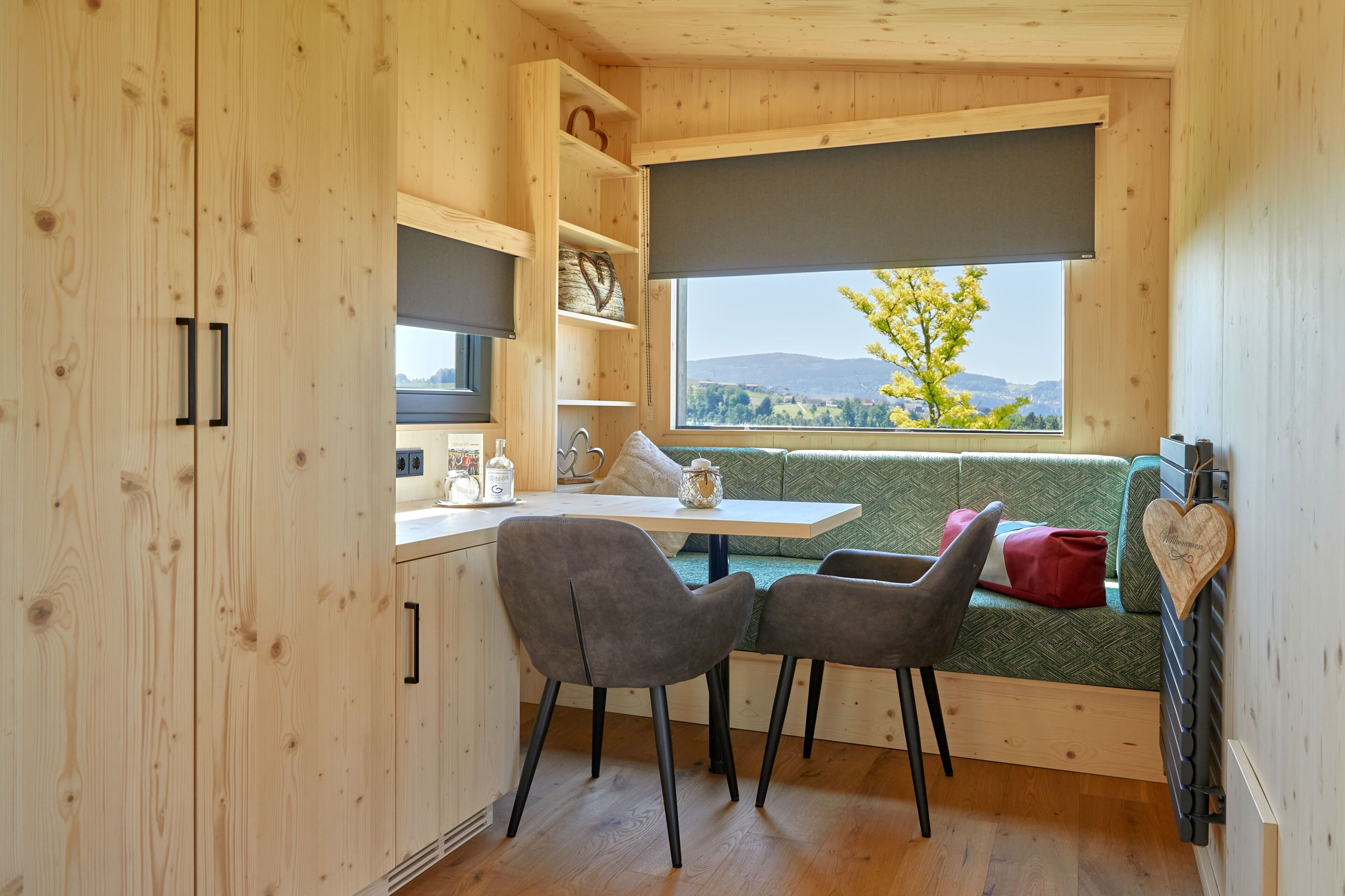 A compact kitchen with a dining area, a window with a view of greenery and hills, and a built-in bench with cushions.