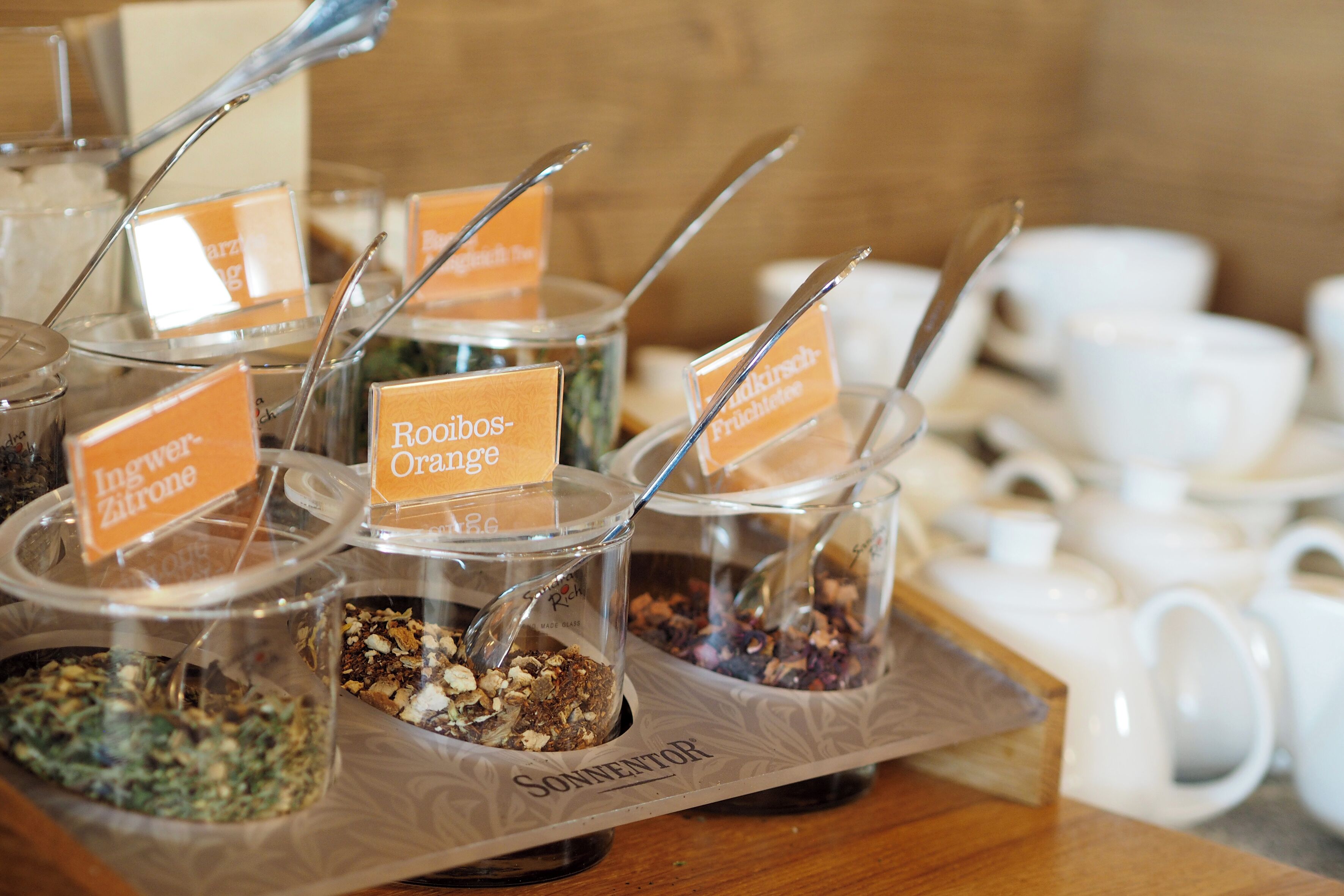 Disposição de buffet com recipientes etiquetados contendo diversos alimentos e utensílios.