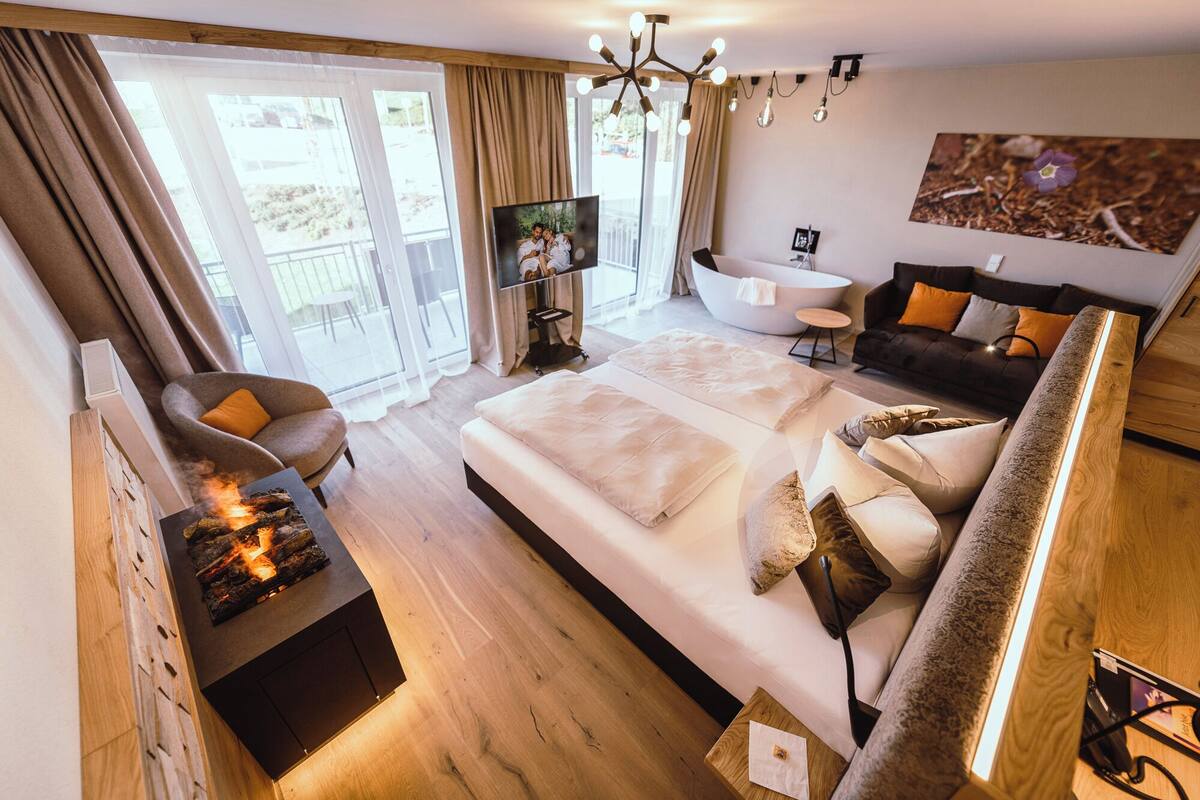 A modern hotel room with a large bed, a sofa, a TV, and a fireplace.