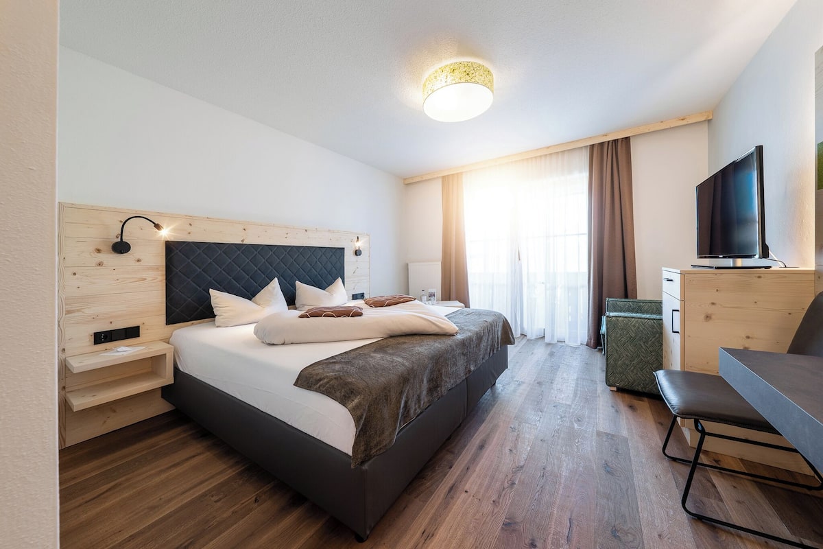 A modern hotel room with a large bed, a desk, a television, and a wooden headboard.
