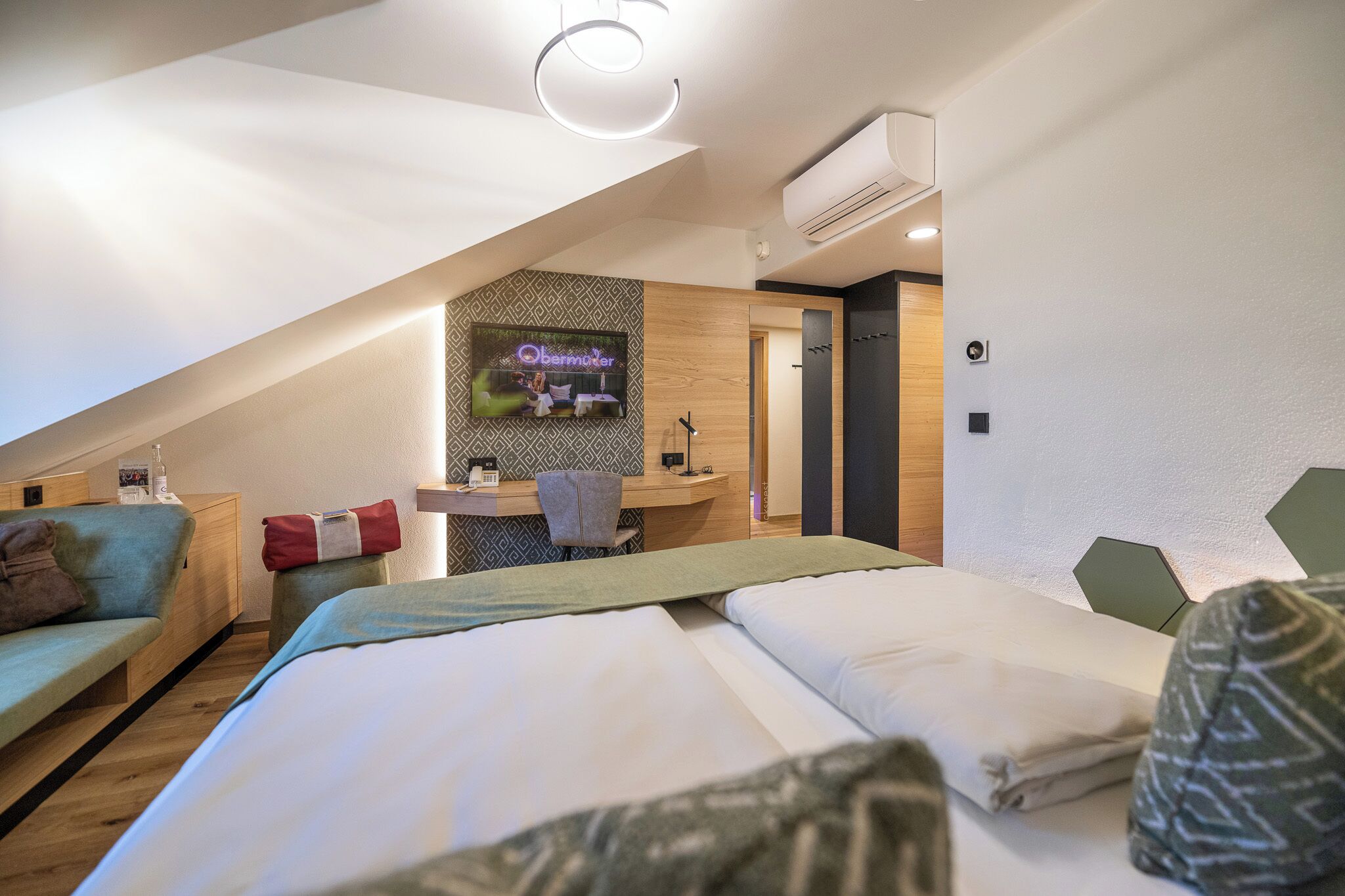 A modern hotel room with a large bed, a desk, and a TV displaying 'Obermutter'.
