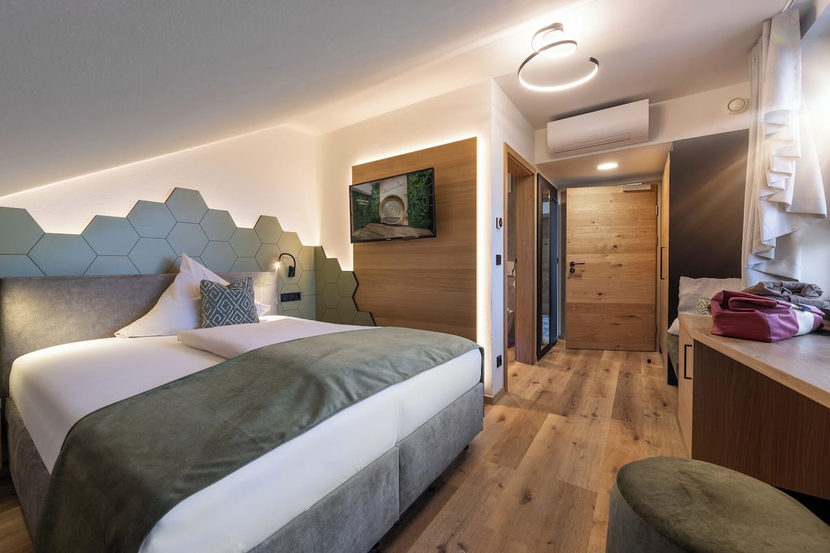 A modern hotel room with a large bed, a wooden headboard, a mounted TV, and a wooden door leading to a bathroom.