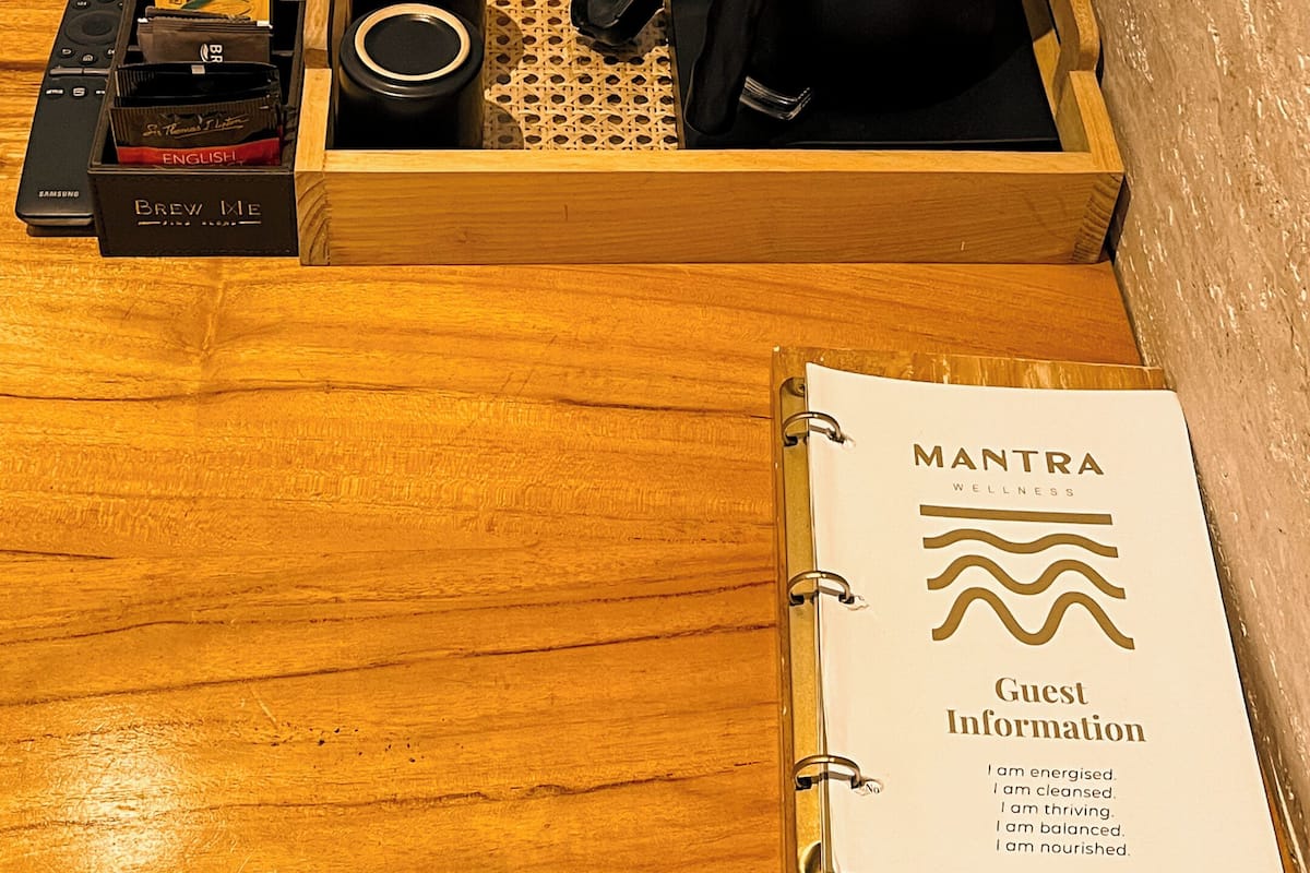 A wooden tray with a coffee maker, cups, a remote control, and a booklet titled 'MANTRA WELLNESS' on a wooden table.