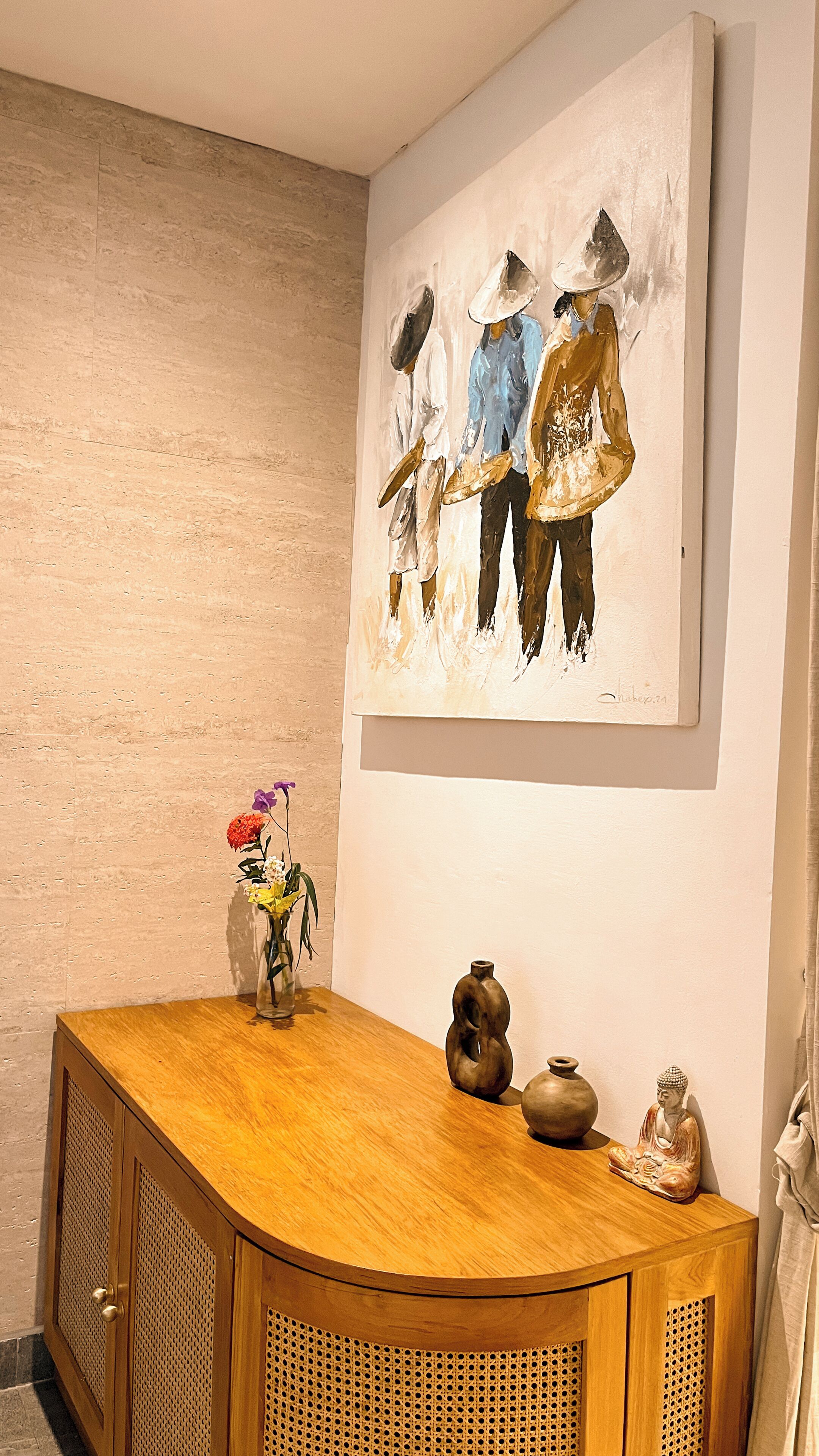 A wooden cabinet with a vase of flowers, a Buddha statue, and a painting on the wall.
