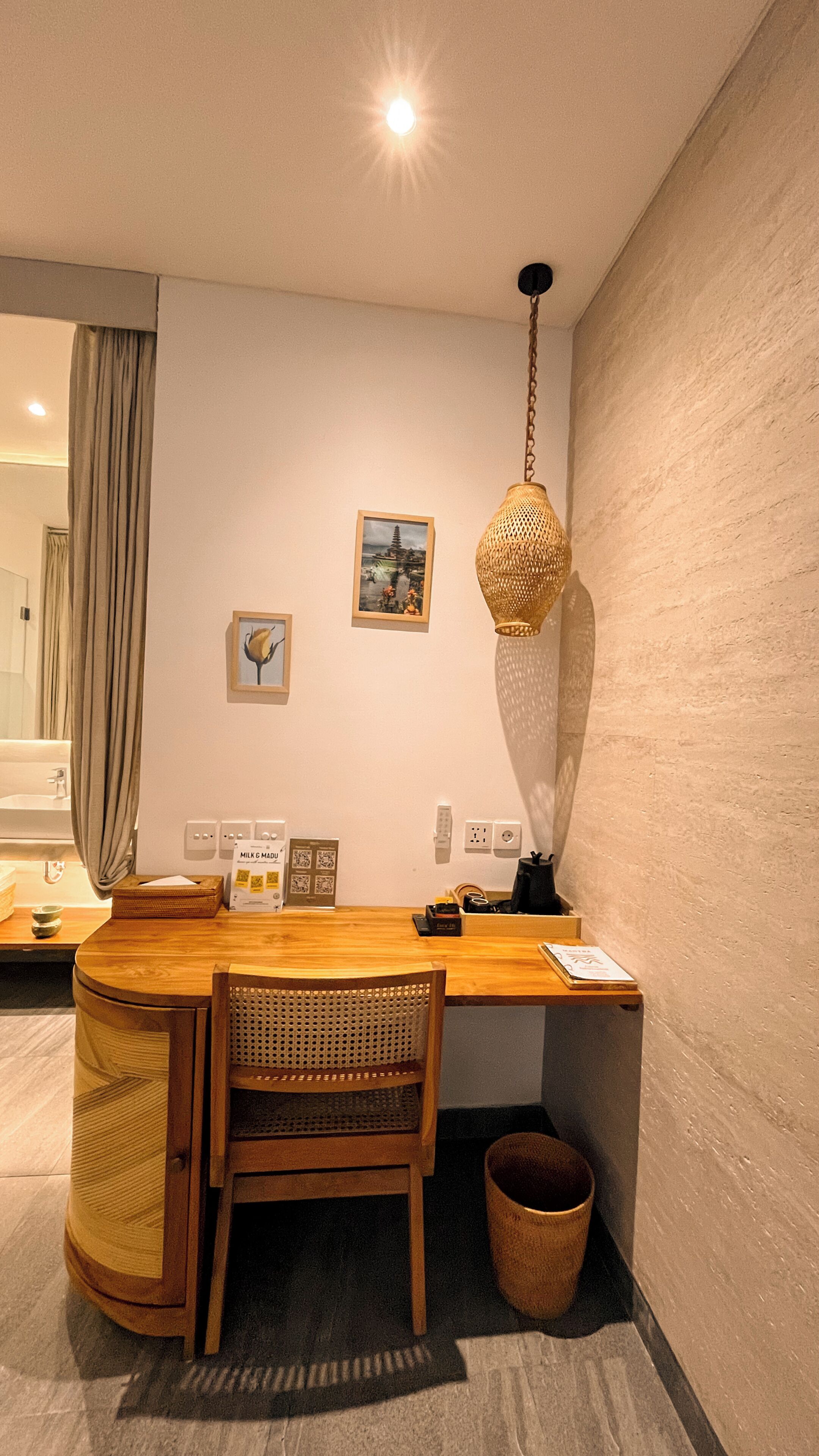 A modern room with a wooden desk, a woven lamp, a chair, a small trash bin, and a wall-mounted picture.