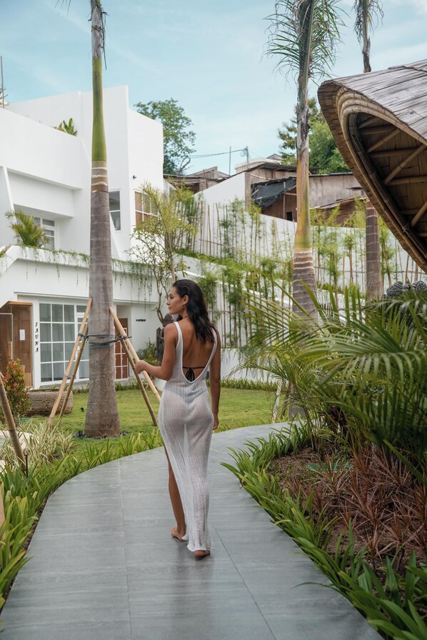 Una mujer caminando por un sendero empedrado en un jardín con palmeras y un edificio moderno al fondo.