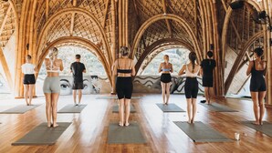 A group of people standing on yoga mats in a room with a bamboo ceiling and a mural of a face on the wall. - Mantra Wellness Centre (Pecatu)