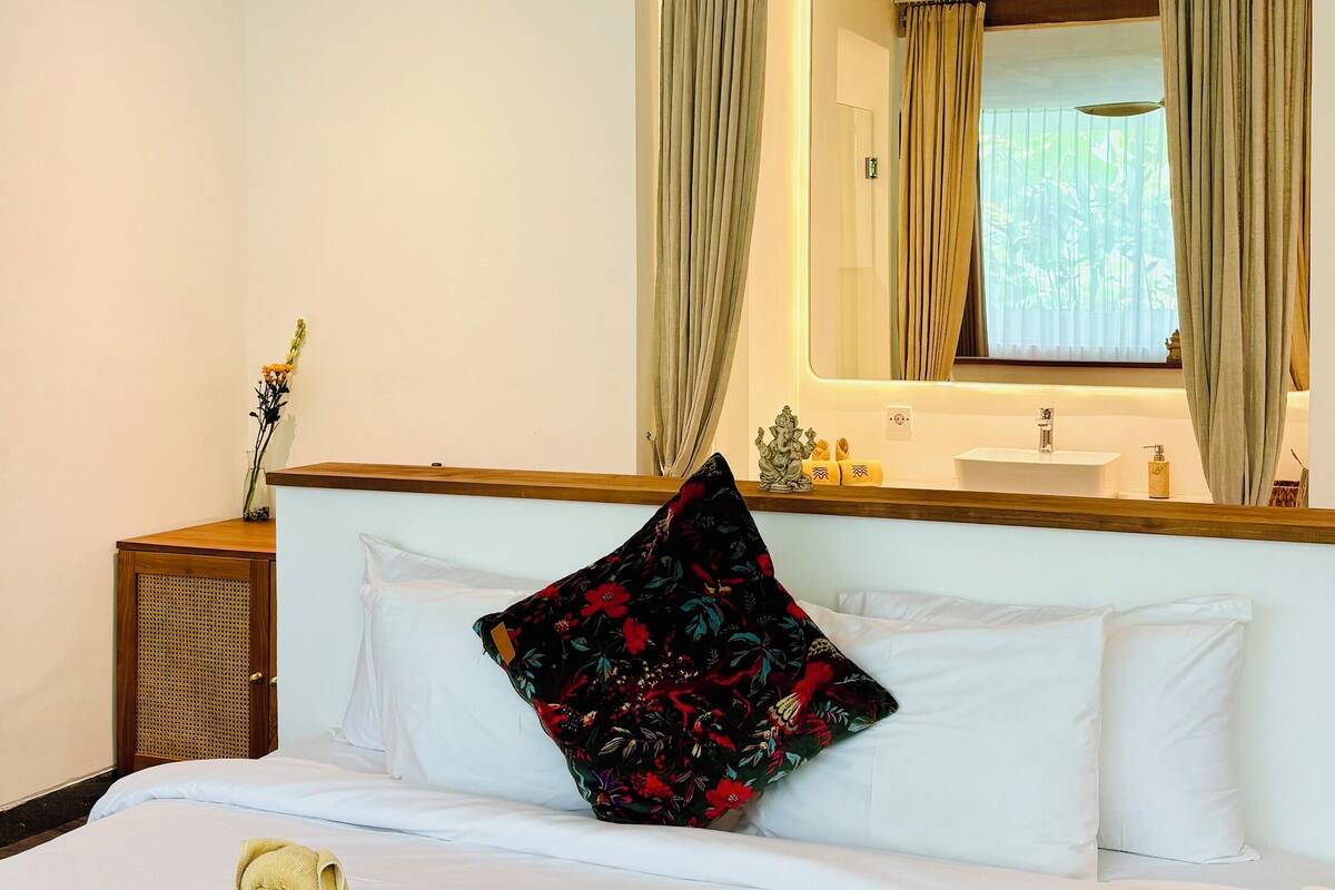 A hotel room with a bed, a green throw blanket, a floral pillow, a wooden nightstand, a mirror, and a ceiling fan.