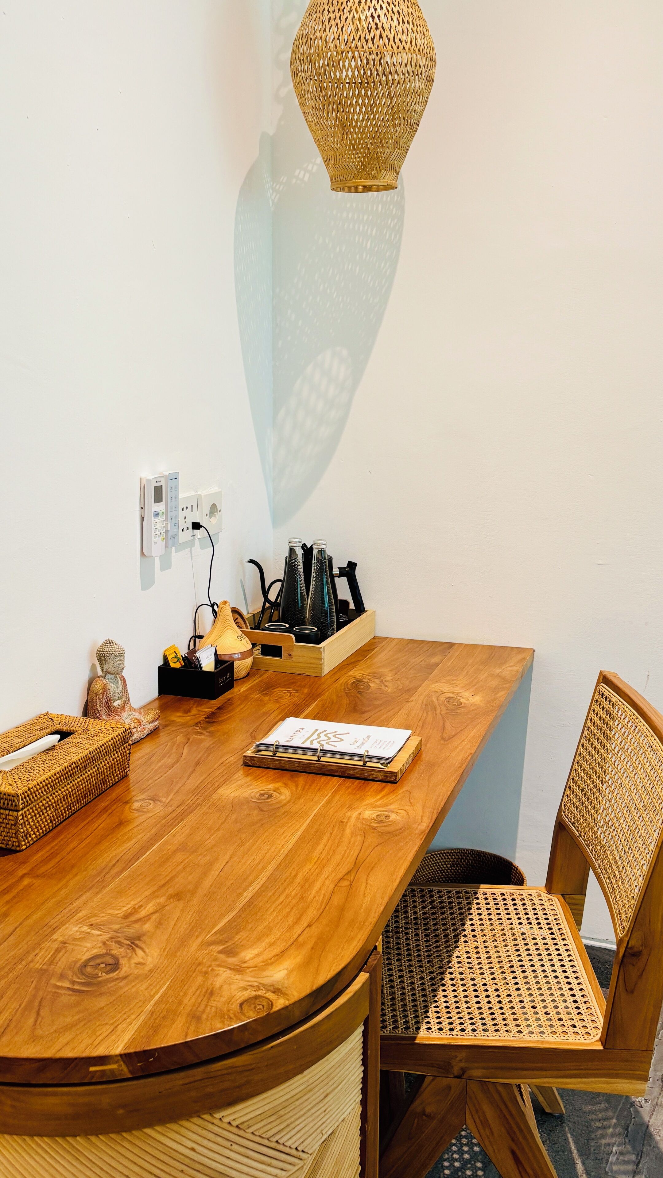 Une table de salle à manger en bois, accompagnée d’une chaise en osier, d’une petite statue de Bouddha, d’une boîte de mouchoirs, d’un carnet et d’un porte-bouteille.