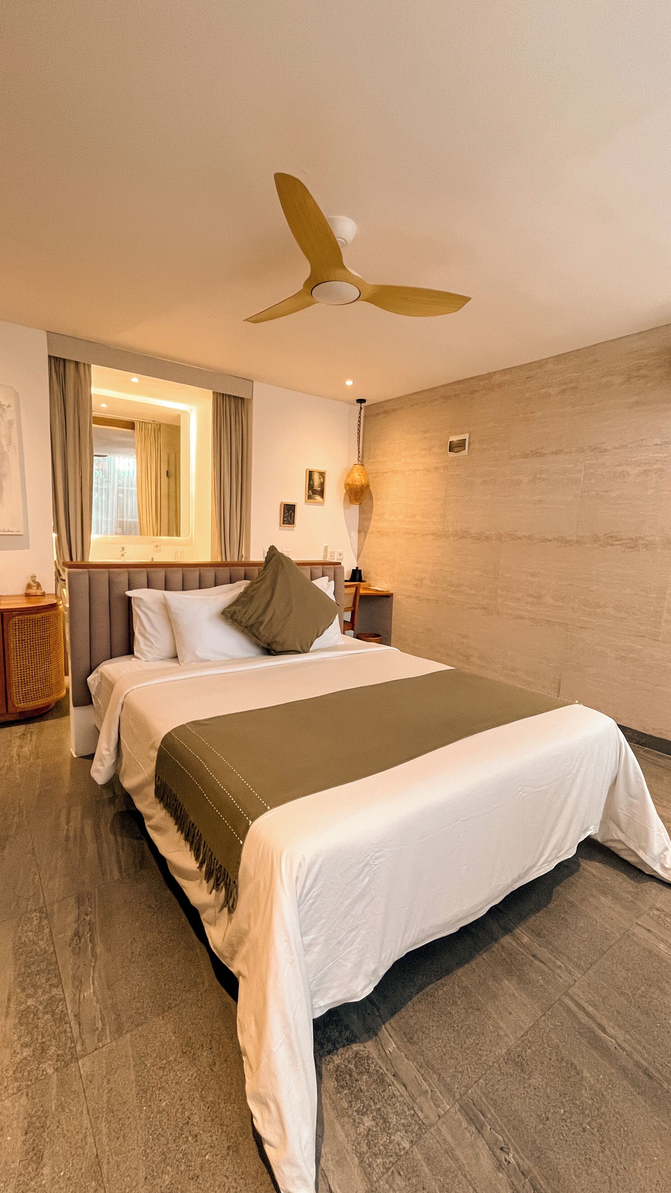 A hotel room with a bed, a ceiling fan, a bedside table with a lamp, and a view of the outside through a window.