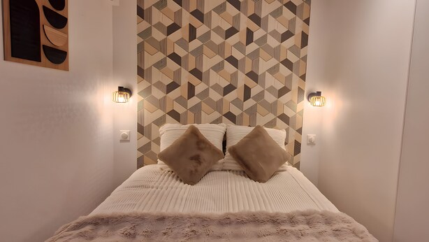A bedroom with a geometric-patterned headboard, two wall-mounted lights, and a fluffy white bedspread. - Maison Bonhage (Chaumont)